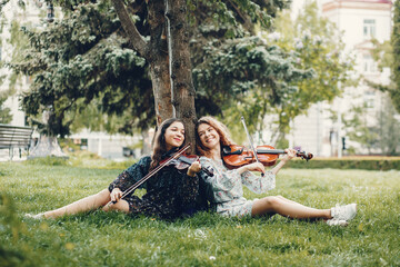 Girls in a summer park. Two ladies with a violin. Friends sitting on a grass