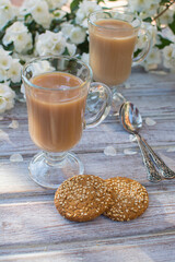 Two glass glasses with coffee and two oatmeal cookies with sesame seeds on a wooden table against the background of blooming jasmine.
