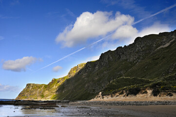 Naklejka premium Dunes de sable et herbe verdoyante sur une plage du littoral au nord de l'Irlande. Ciel bleu et nuages blancs contrastant avec les couleurs des falaises.