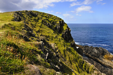 Fototapeta na wymiar falaise couverte de verdure et rochers sur lesquels la mer bleue indigo vient s'abattre formant une écume blanche, ciel bleu et nuages blancs au nord de l'Irlande.