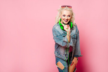 Happy stylish blond woman in pink glasses with big headphones listening music and dancing on colored background