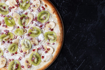 Fruit pizza with pomegranate berries, slices of kiwi and banana on a dark background close-up