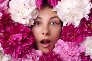 Attractive woman with opened mouth mouth, with astonished facial expression, female with beautiful eyes looks shocked, girl surrounded with white, pink and burgundy peony flowers.