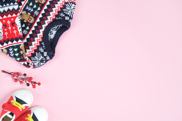 Christmas sweater, baby shoes and holly twig on pink background. Copy space. Christmas clothes flatlay.