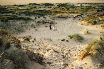 Happy wedding pair walking in sand dunes. Barefoot bride and groom holding hands walking in sand in evening light. Wedding day concept.