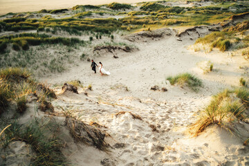 Happy wedding pair walking in sand dunes. Barefoot bride and groom holding hands walking in sand in evening light. Wedding day concept.