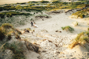 Happy wedding pair walking in sand dunes. Barefoot bride and groom holding hands walking in sand in evening light. Wedding day concept.