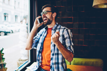 Happy young man talking on smartphone and rejoicing of victory of successful project.Excited hipster guy celebrating win calling on cellular.Amazed blogger received good news during phone conversation