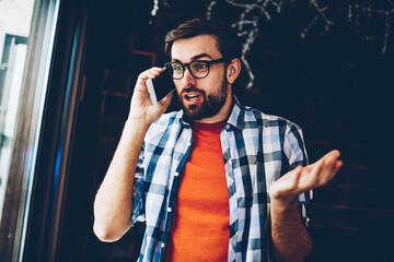 Amazed bearded young man surprised with news talking with friend during mobile conversation on digital smartphone device.Wondered hipster guy excited with shocked story calling on modern telephone