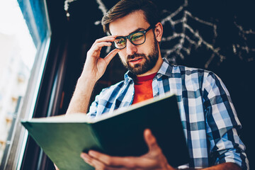 Concentrated smart young man with beard and optical eyeglasses searching useful information in textbook for coursework.Pensive hipster guy reading book preparing for studying seminar in coworking