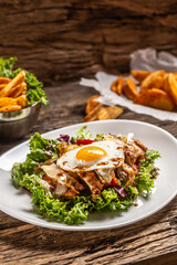 Chicken salad with sunny side up egg and sauce on a white plate and rustic wood, with potato wedges on a side