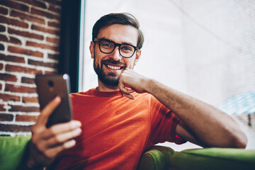 Portrait of successful bearded male blogger laughing at camera while downloading app on smartphone device via free 4G internet connection.Positive hipster guy making payment online on cellular