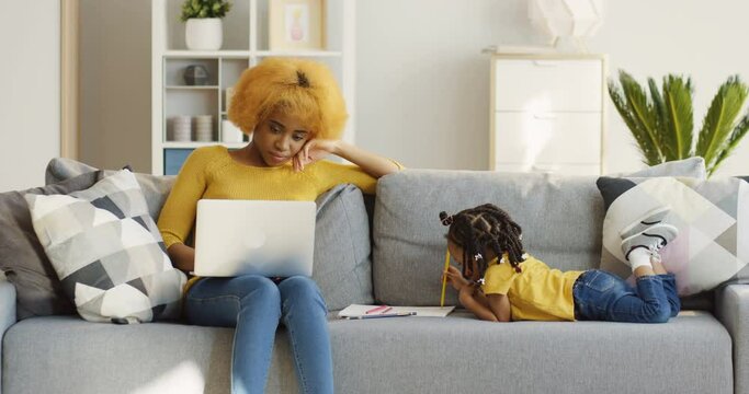 Young African American good looking woman working on the laptop computer and thinking while siting on the gray sofa in the living room and her cute little daughter drawing . Indoors
