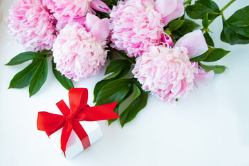 A bouquet of pink peonies, a box with a gift tied with a red bow on a white background. Happy birthday or wedding day greeting card.