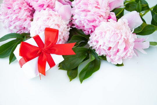 A bouquet of pink peonies, a box with a gift tied with a red bow on a white background. Happy birthday or wedding day greeting card.