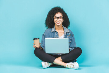 Naklejka premium Portrait of smiling young afro american woman using laptop while sitting on a floor with legs crossed isolated over blue background. Drinking coffee or tea.