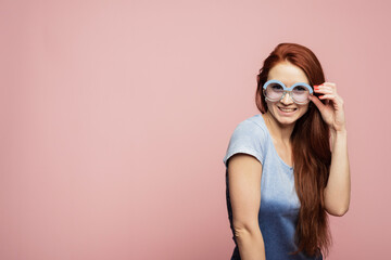 Naklejka premium Indoor portrait of redhead lady in sunglasses fooling around in studio. Beautiful girl in blue gradient shirt wearing sunglasses posing, smiling on pink background in studio