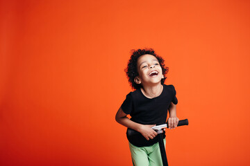 Curly joyfully laughing boy riding on scooter on his hands and head on an orange background in studio