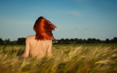 Red-haired topless girl stands in a grain field with her back turned away.