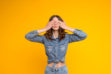 Calm beautiful curly brunette young woman in blue denim casual shirt and jeans covering her eyes with both hands,standing isolated on yellow background.I am afraid,shy,blind