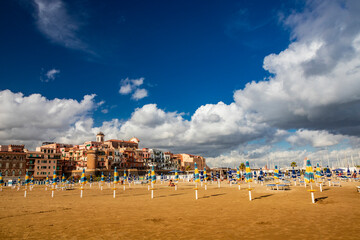 The closed umbrellas, the empty sunbeds and deckchairs and the deserted beach in a bathhouse, on a windy day in late summer. The cloudy blue sky. Nobody, no people. Nettuno, Rome, Lazio, Italy.