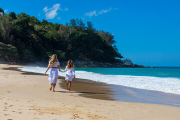 Young beautiful mother with her charming daughter in white clothes walk on a sandy beach near the water