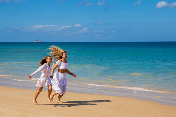 Young beautiful mother with her charming daughter in white clothes walk on a sandy beach near the water