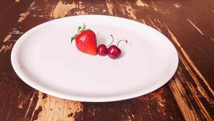 Top view of red strawberry and two sweet red cherries in the white plate on a wooden background. Space for text. Design concept.
