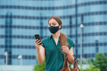 A young woman in a navy blue medical face mask to avoid the spread of coronavirus holding her leather bag in the center of the city. A girl with long hair reading the news on a smartphone in downtown.