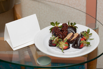 
On a glass table is a white plate with chocolate strawberries, next to a plate is a white card for...