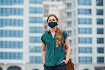 A portrait of a woman in a medical face mask is thrusting hands into pockets of trousers while walking in the center of the city. A girl in a protective mask keeping social distance on the street