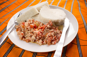 A white plate of boiled yam and fried scrambled eggs prepared with sliced onions, tomatoes and red pepper. Ready to eat and served on a colorful African pattern background with a knife and fork 