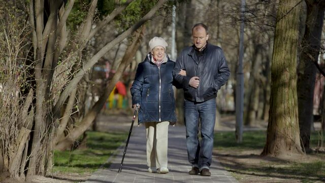 An elderly woman walks with her son enjoying an evening walk in park, talking and smiling. Man helps an elderly woman to walk. Couple of two generations - mother 81 years old and son 43 years old.