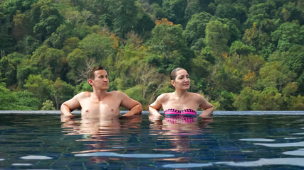 smiling couple relaxes in large hotel swimming pool blue water against green hilly landscape under bright summer sunlight
