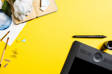 top view of drawing tablet with blank screen, clips, stylus, painting, notebook and flower on yellow