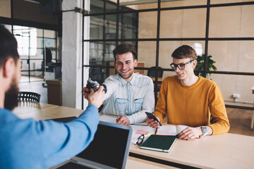 Successful male colleagues enjoying work time together sitting at desktop indoors while one of them messaging via smartphone, smiling hipster guy taking retro camera from friend on front with laptop