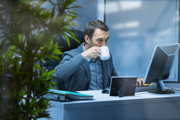 The Manager is sitting at an office Desk, typing on a computer and drinking a hot drink.