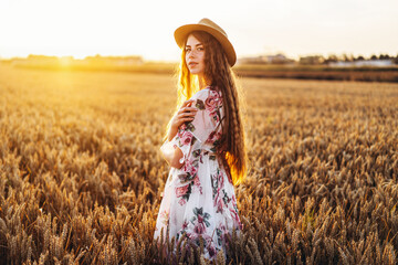 Portrait of a beautiful young woman with curly hair and freckles face. Woman in dress and hat posing in wheat field at sunset and looking at camera © bedya