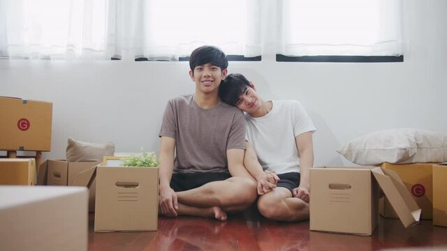 Portrait of young asian couple man sitting, smiling and looking to camera with carton package box storage to move in empty new home. LGBTQI relationship and relocating a new house concept