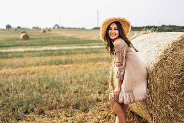 Beautiful young girl with long hair in sunnglasses and straw hat posing on a wheat field near hay...