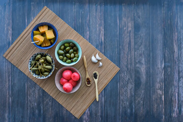 Assorted pickles: aubergines, olives, spring onions and capers in bowls on a wooden surface, top view, copy space.