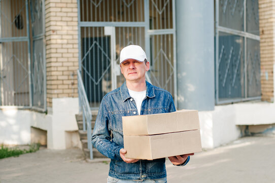 male courier in the blue costume and a cap taking out mail carton boxes from the white van on the sunny day in the street. Outdoor.