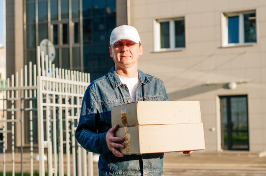 male courier in the blue costume and a cap taking out mail carton boxes from the white van on the sunny day in the street. Outdoor.