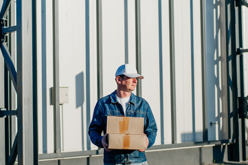 male courier in the blue costume and a cap taking out mail carton boxes from the white van on the sunny day in the street. Outdoor. - obrazy, fototapety, plakaty