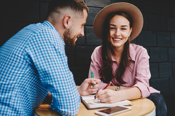 Portrait of happy young woman in hat smiling at camera while spending leisure time together with boyfriend in stylish cafe interior.Positive girlfriend smiling at camera while writing list to do