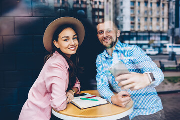 Romantic couple sitting at table in cafe during date posing for selfie on smartphone camera,...