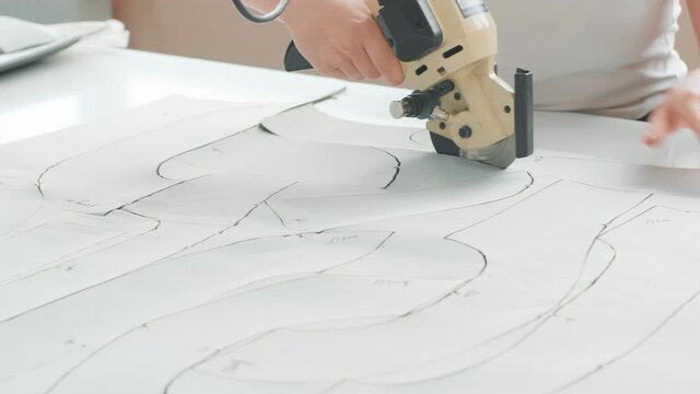 Female worker on a sewing manufacture uses electric cutting fabric machine. Sewing production line