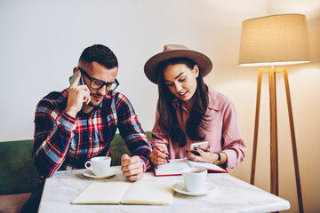 Male and female colleagues busy while making research for startup project talking on phone and writing in notebook,businessman making banking during mobile conversation cooperating with partner
