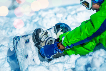 Close up of a boy attach snowboard to the legs sitting in snow about to start going downhill on ski...