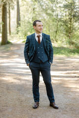 man in a dark blue suit and in a red tie posing in the forest.